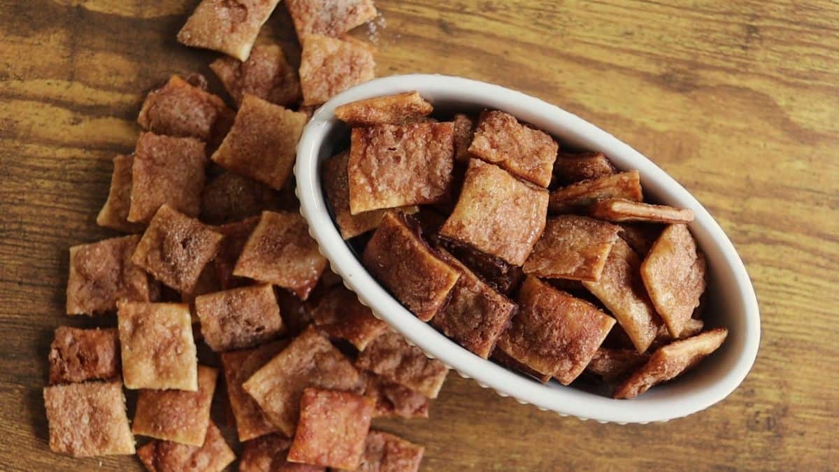 A white oval dish filled with small, square cinnamon sugar crackers sits on a wooden surface, with more crackers scattered beside it.