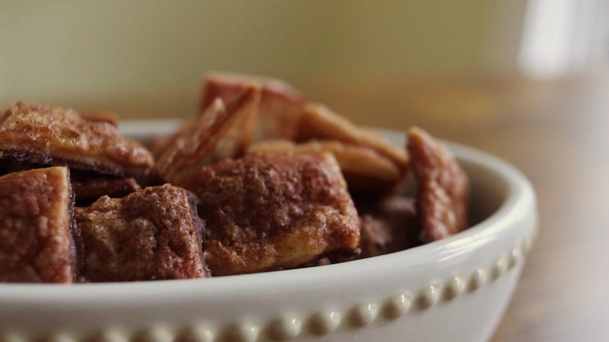 A close-up of a white bowl filled with cinnamon sugar-coated baked chips or pastry pieces, sitting on a wooden surface with a softly blurred background.