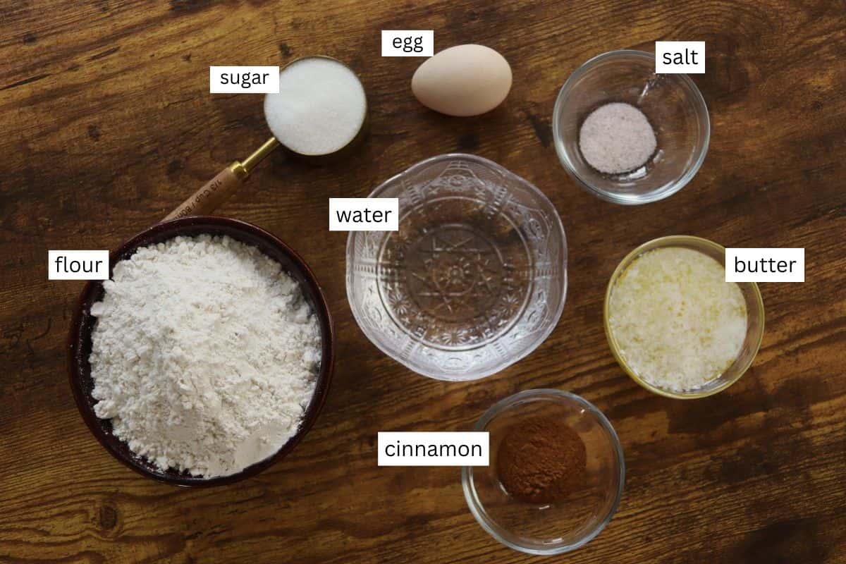 Bowls and cups with labeled baking ingredients on a wooden surface: flour, sugar, water, egg, salt, melted butter, and cinnamon.