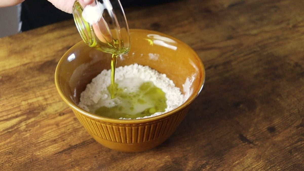 Oil being poured from a small dish into a bowl of flour that is sitting on a wooden surface.