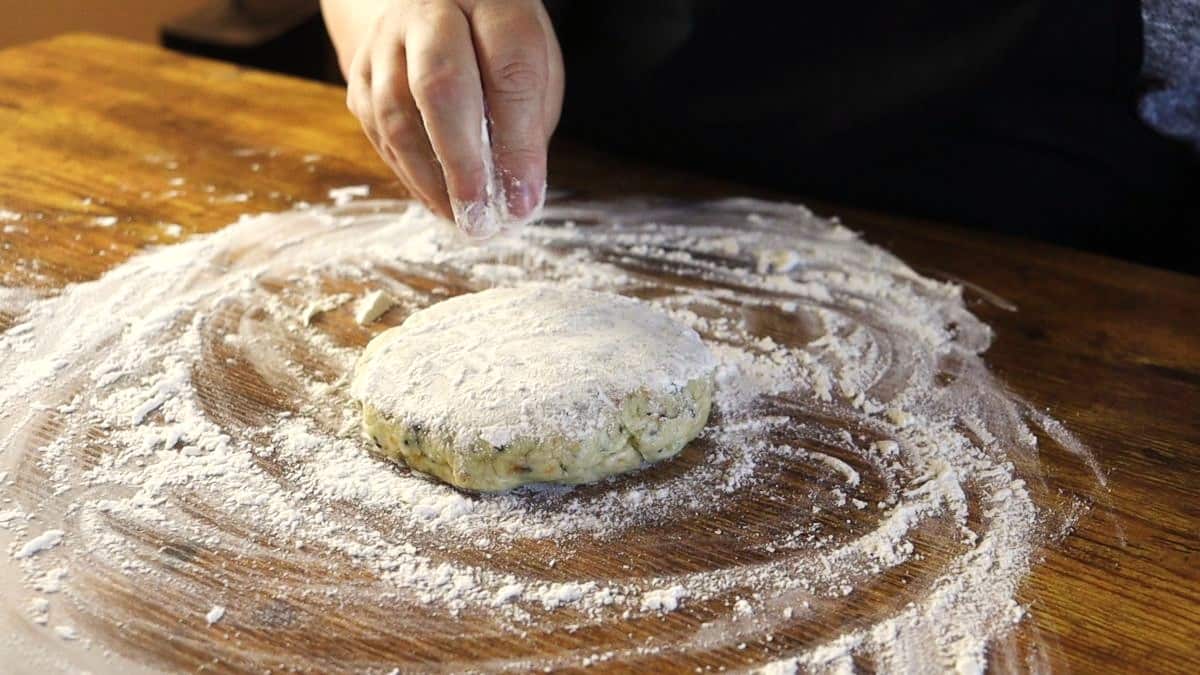 Hand sprinkling flour over a disk of dough sitting on a floured wooden surface.