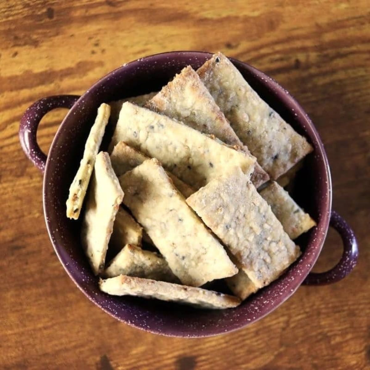 Rectangular crackers with seeds visible in them in a purple bowl with handles sitting on a wooden table.