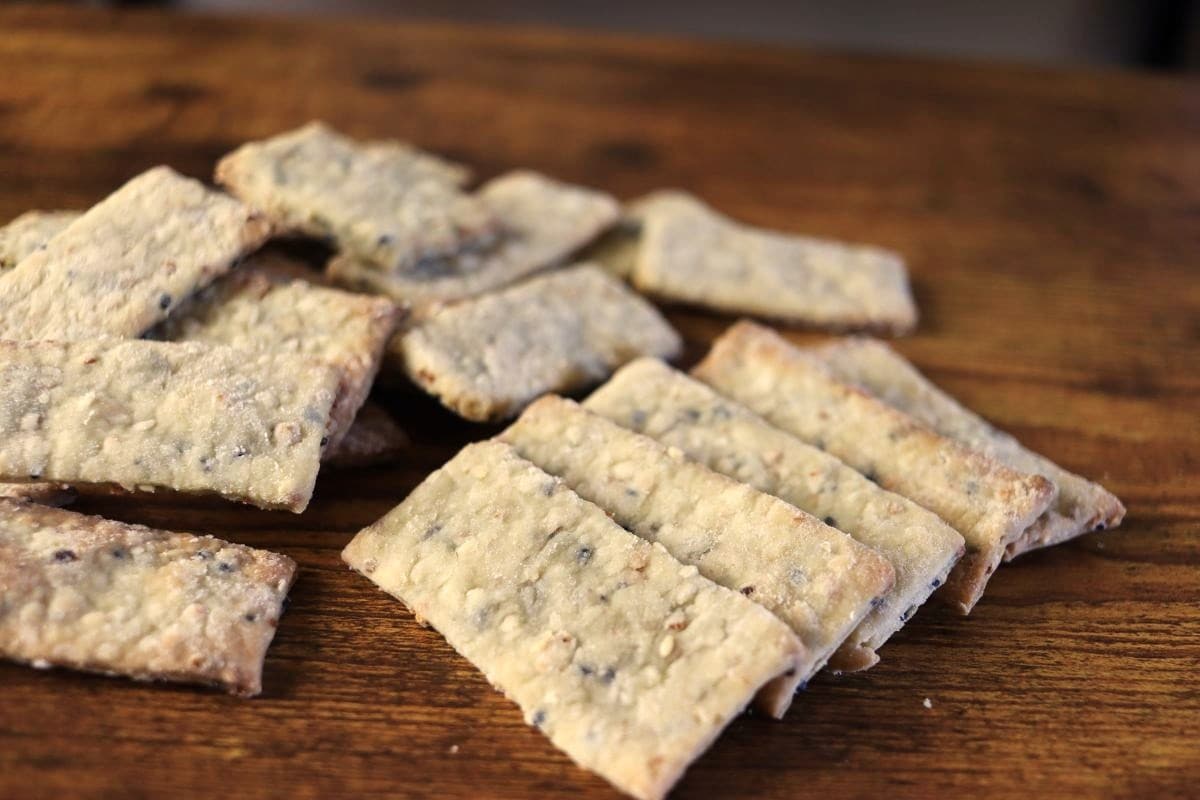 A pile of rectangular, homemade crackers with visible seeds and grains sits on a wooden surface. The crackers appear crispy and lightly browned.