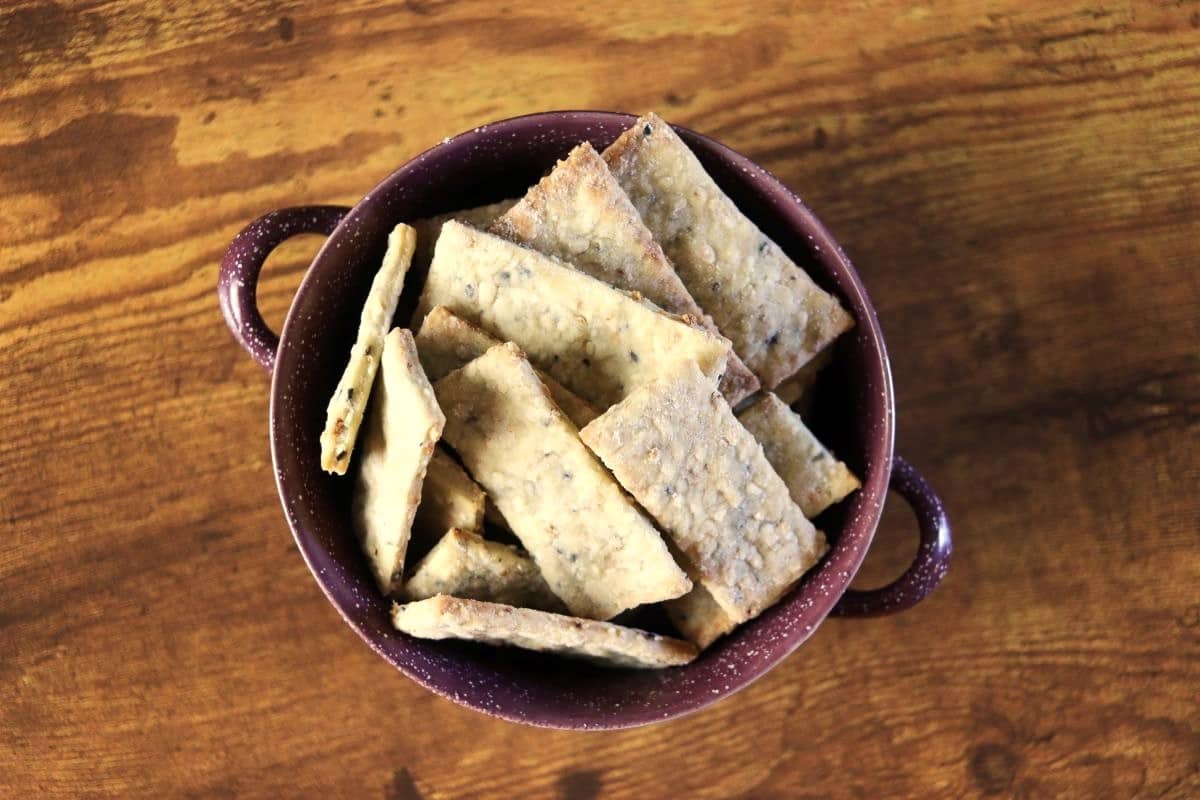 A purple bowl with handles filled with rectangular, homemade crackers sits on a wooden surface. The crackers appear crisp and speckled with seeds.