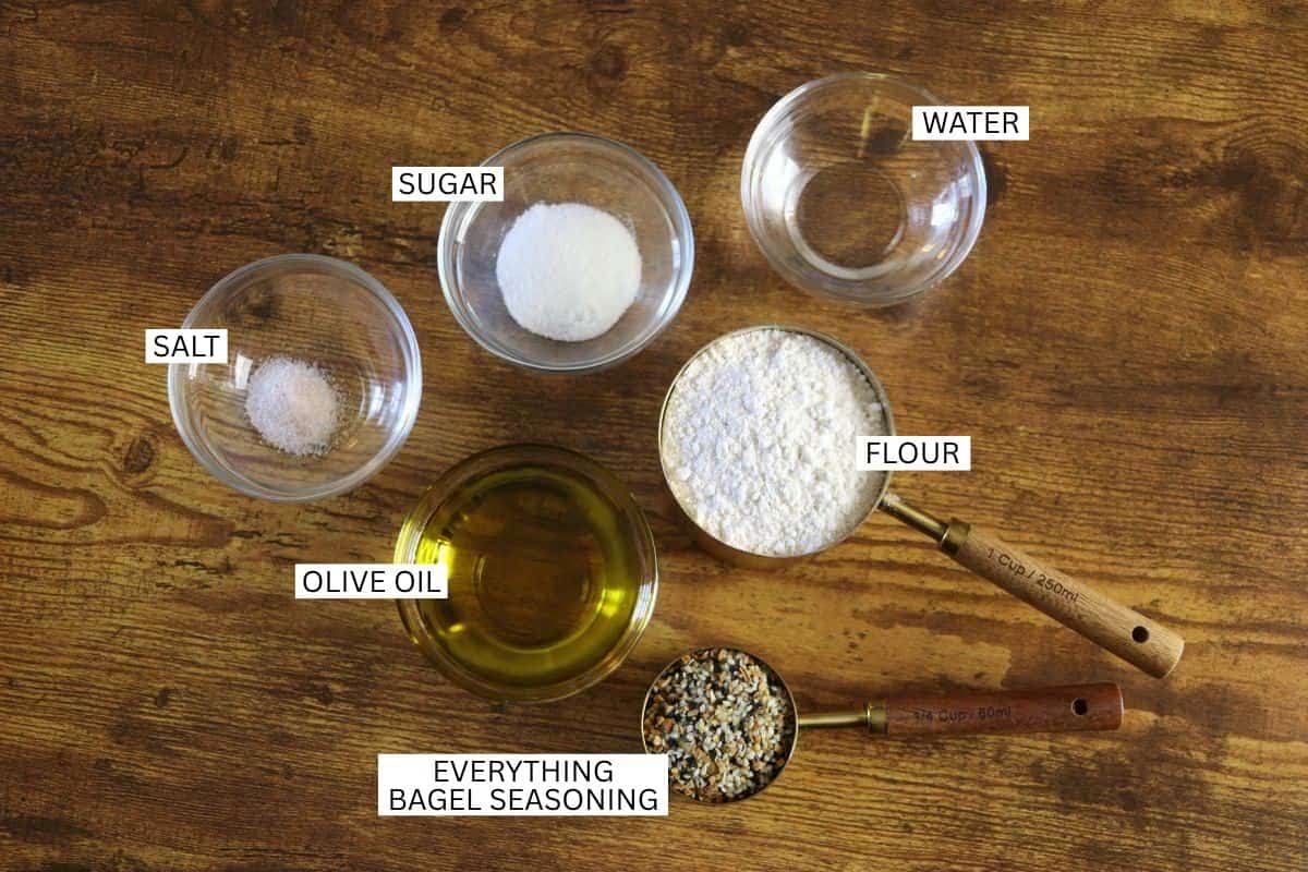 Five labeled ingredients on a wooden surface: sugar, salt, water, flour, olive oil, and a measuring cup with everything bagel seasoning.