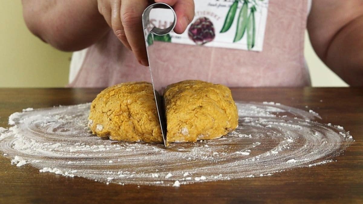 Woman using a bench press to cut an orange ball of dough sitting on a floured wooden surface in half.