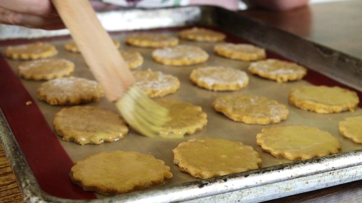 Basting egg mixture over orange dough shapes on a baking sheet with a wooden basting brush.