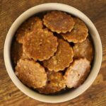 Round, glossy finished, brown crackers in a white bowl sitting on a wooden surface.