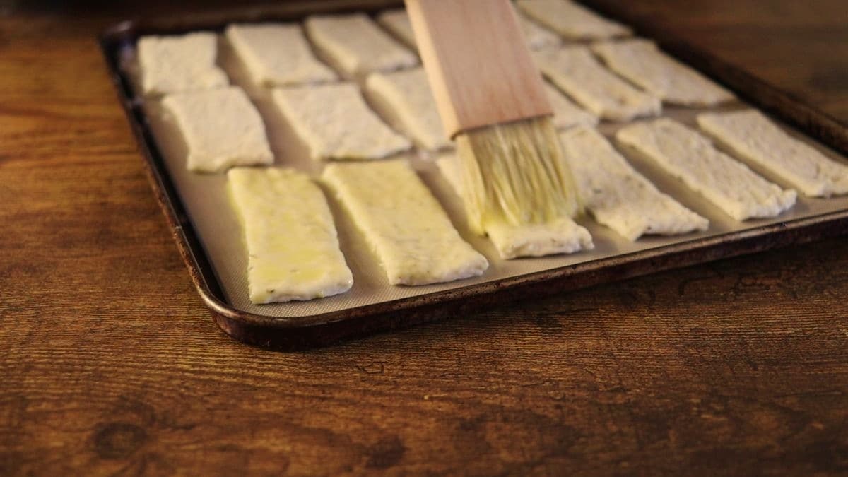 Rectangular pieces of dough on a silicone lined baking sheet being brushed with egg mixture using a basting brush.