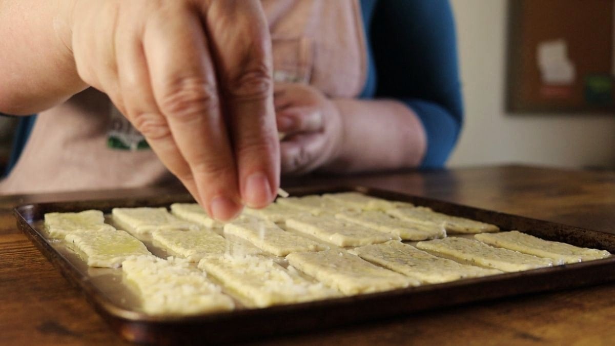 Hand sprinkling cheese over rectangular dough pieces on a baking sheet.