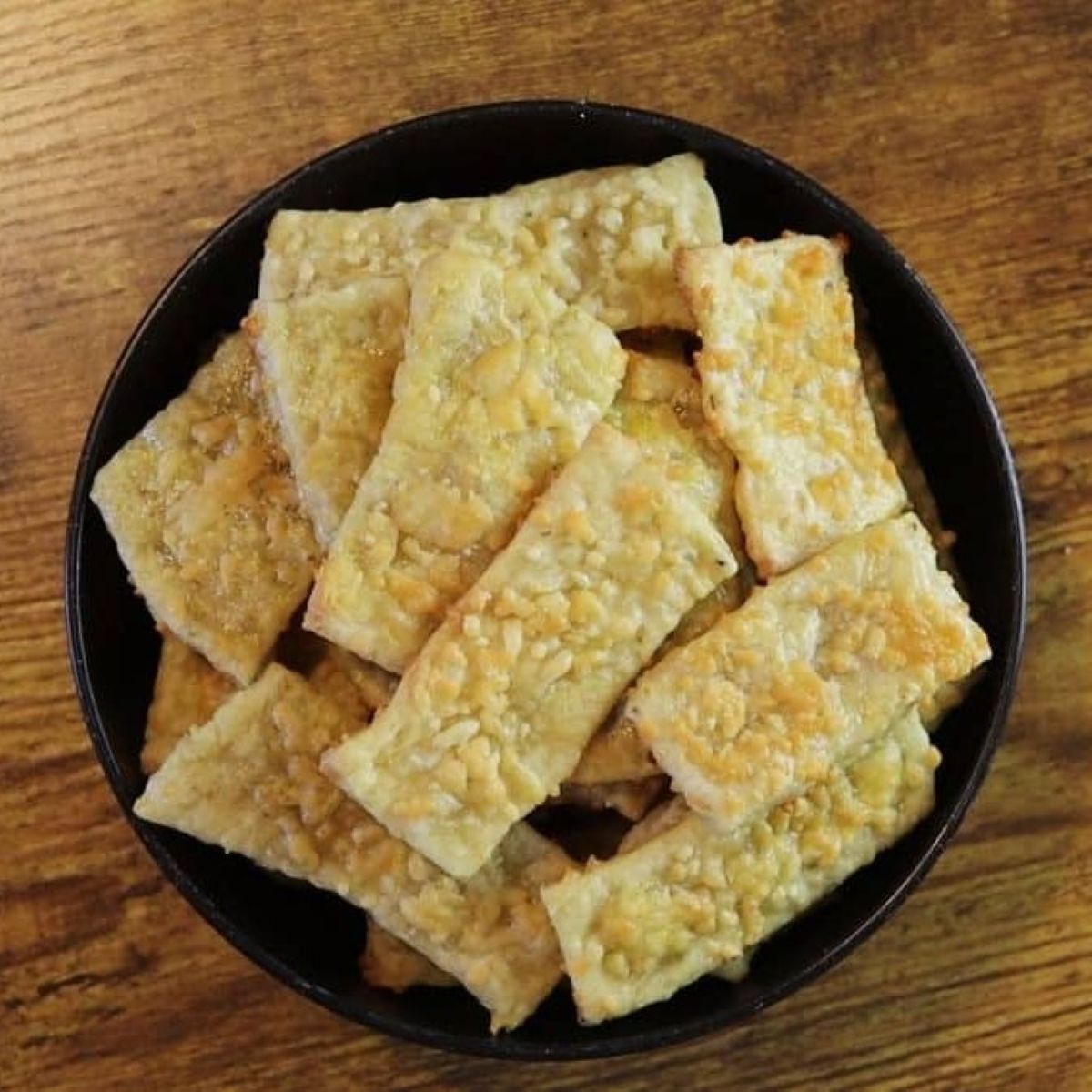 Rustic cheese covered rectangular crackers in a black bowl sitting on a table.