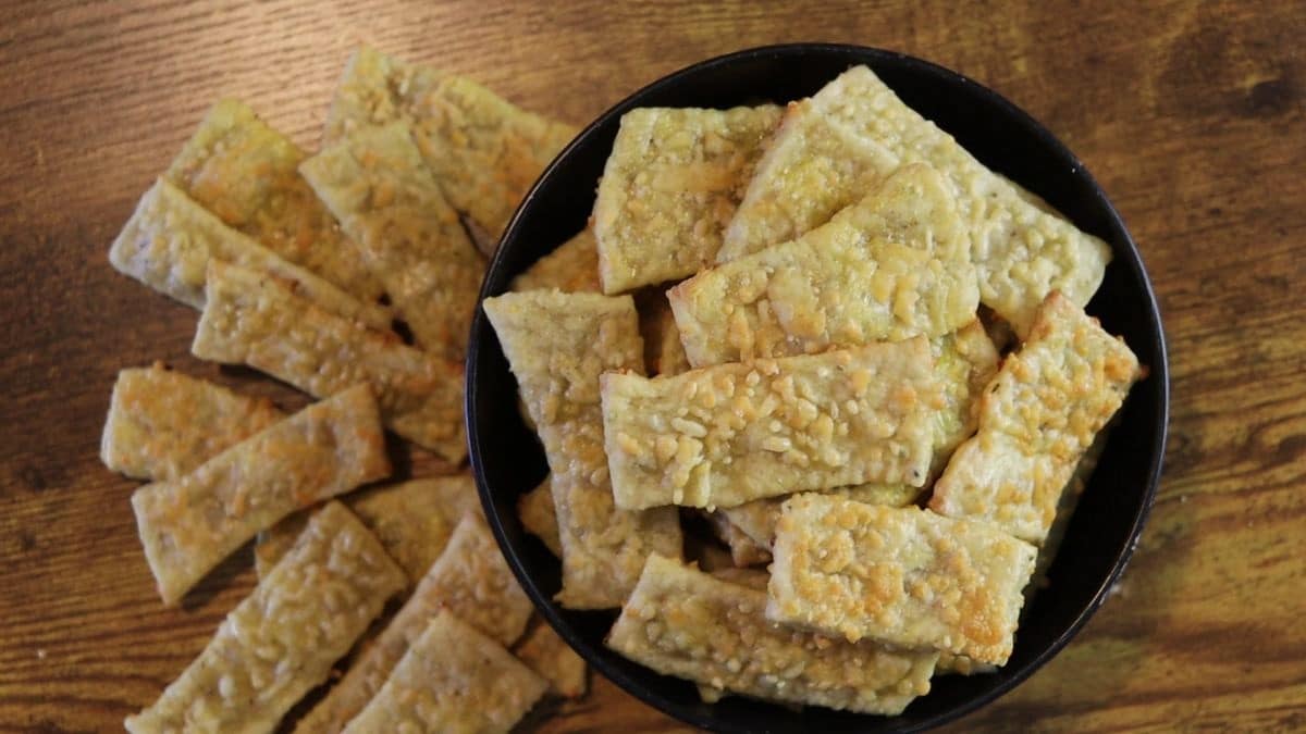 A black bowl filled with rectangular, golden-brown cheese crackers sits on a wooden surface, with more crackers scattered beside the bowl.
