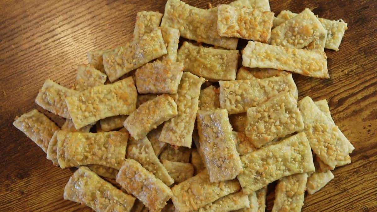 A pile of rectangular, golden-brown cheese crackers with a slightly flaky and textured surface, arranged on a wooden table.