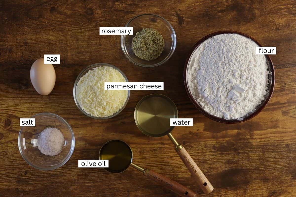Ingredients for a recipe on a wooden surface: a bowl of flour, a bowl of shredded parmesan cheese, a bowl of dried rosemary, a bowl of salt, an egg, a cup of olive oil, and a cup of water. Each is labeled.