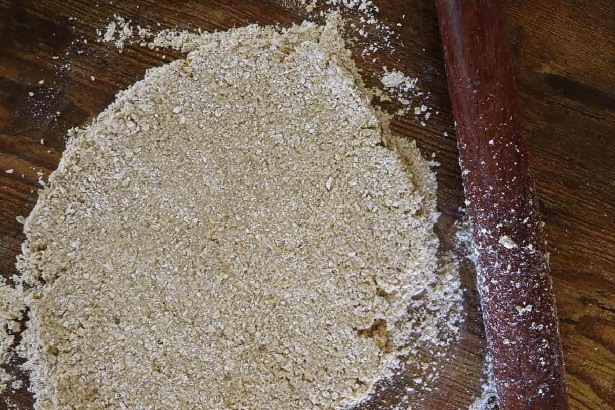 Coarse, grainy dough rolled out on a floured wooden surface with a rolling pin visible to the side of it.