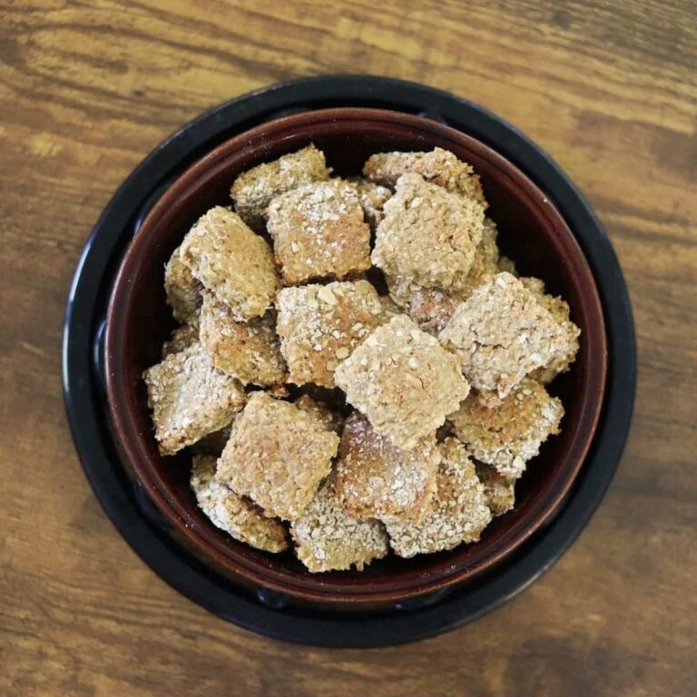 Square golden brown dog treats in a dark bowl sitting on a wooden surface.