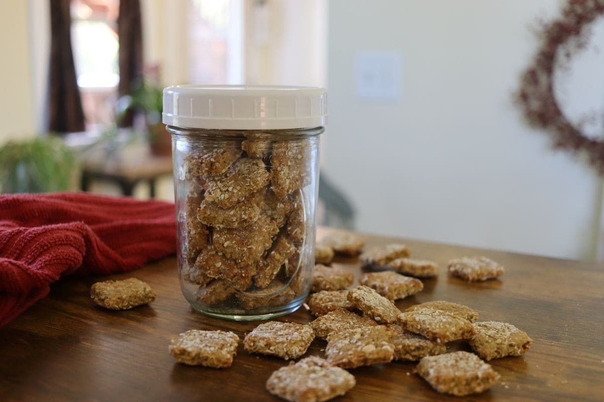 A clear glass jar filled with homemade dog treats sits on a wooden table, with more treats scattered around it. A red cloth is on the table, and a blurred background shows part of a cozy home interior.