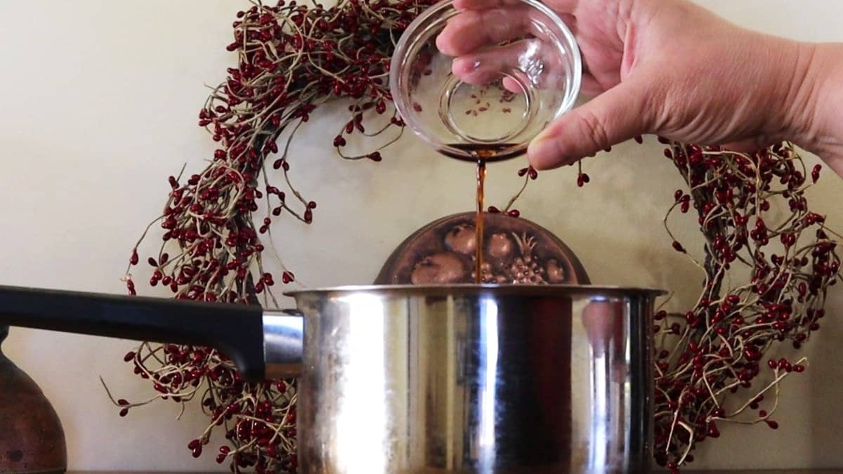 Hand pouring brown liquid from a small glass dish into a saucepan with home decor in the background.
