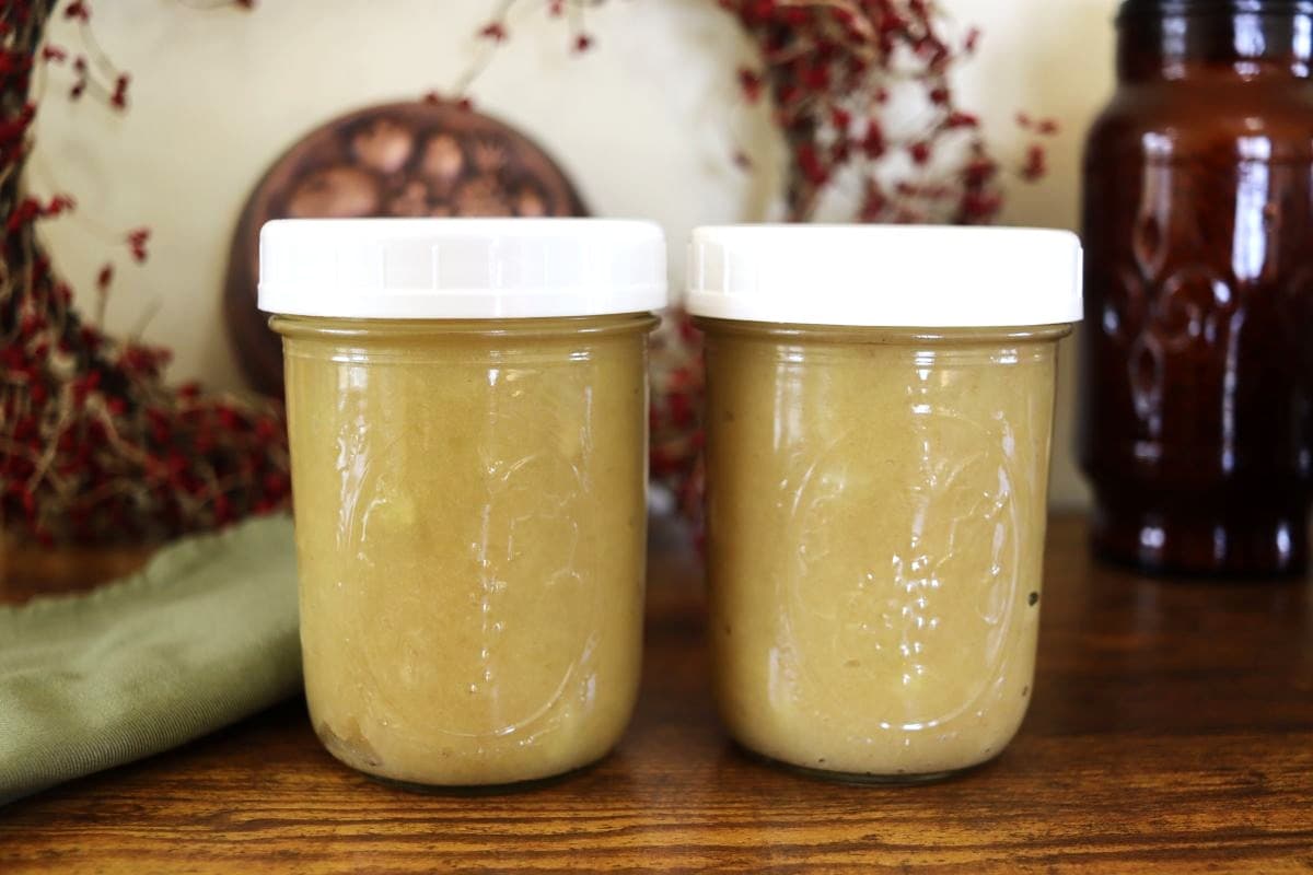 Two mason jars full of homemade applesauce with white lids on top and home decor visible in the background.