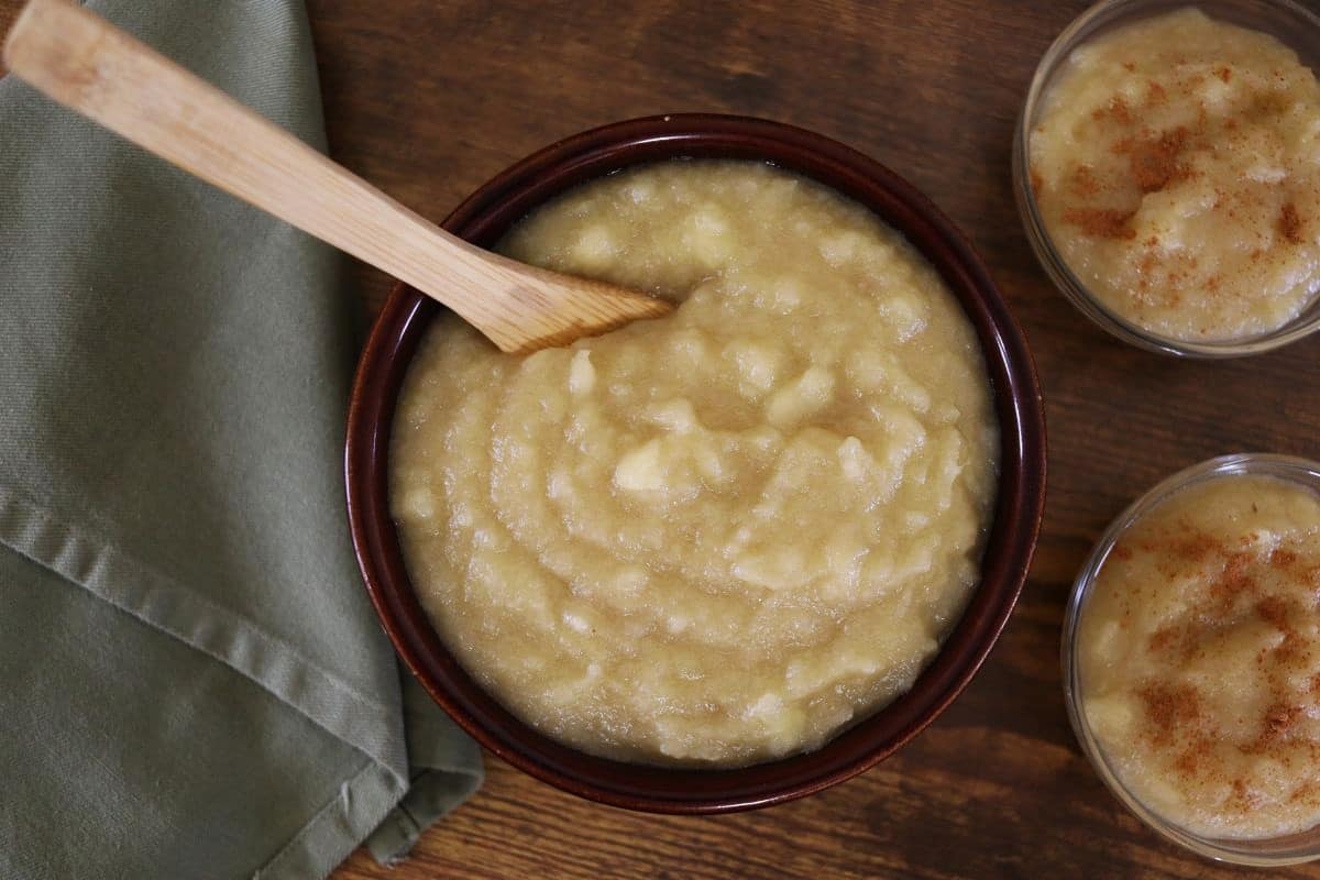 A bowl of chunky applesauce with a wooden spoon, set on a wooden table next to a green cloth napkin. Two smaller dishes of applesauce sprinkled with cinnamon are nearby.