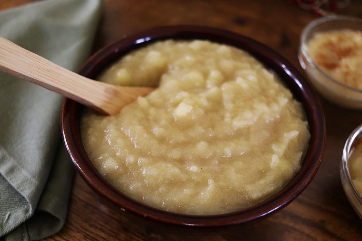 A bowl of chunky applesauce with a wooden spoon, placed on a wooden table next to a folded green cloth napkin and a small dish in the background.