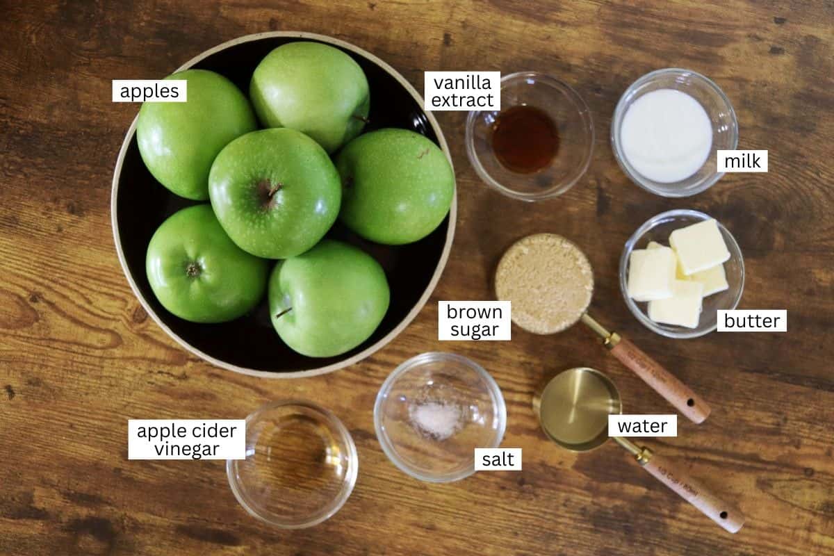 A bowl of green apples and small bowls of vanilla extract, milk, brown sugar, butter, apple cider vinegar, salt, and water are arranged on a wooden surface, each ingredient labeled.