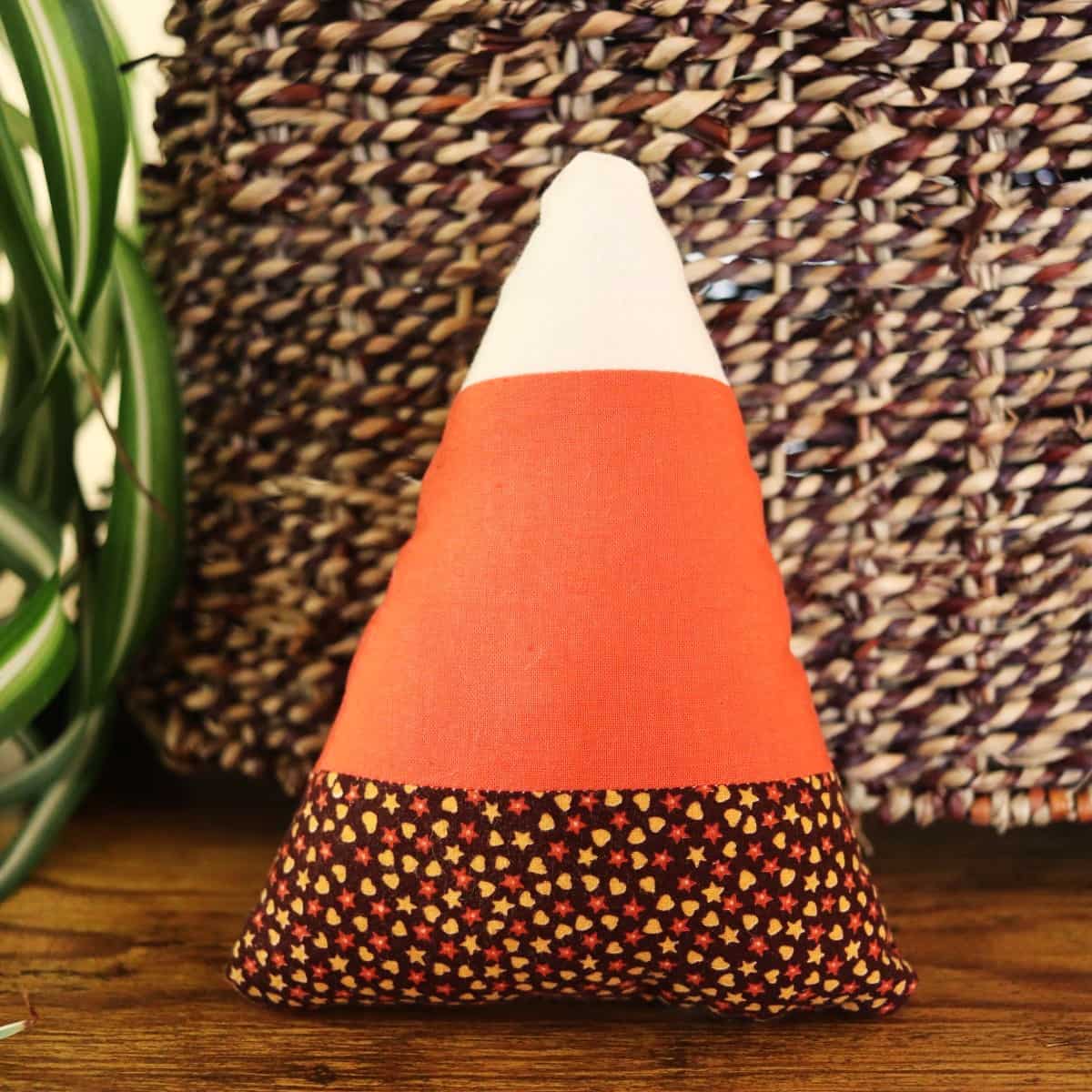 Candy corn shaped stuffie with brown, orange, and white layers standing by a wicker basket with a plant visible to the side.