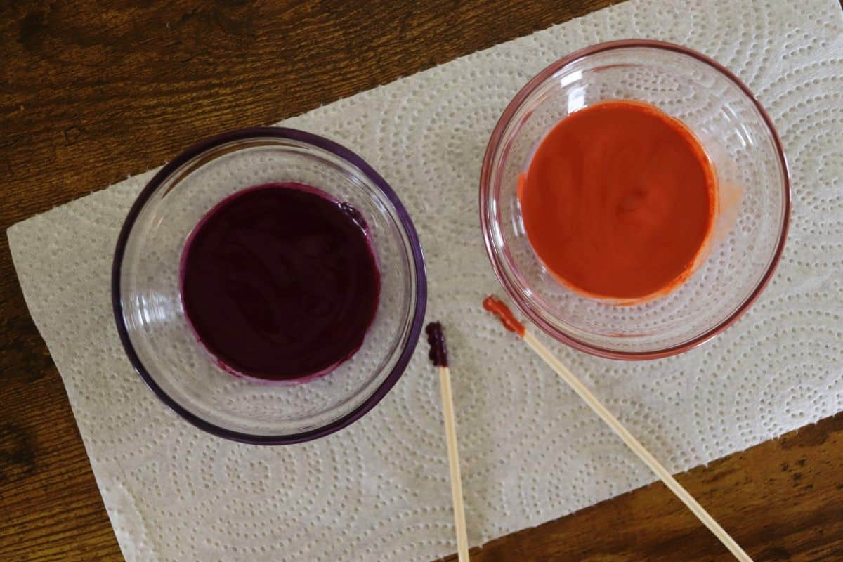 Two small glass dishes sitting on a paper towel. One has purple paint in it. The other has orange paint. There is a wooden dowel next to each dish with paint on the tip.