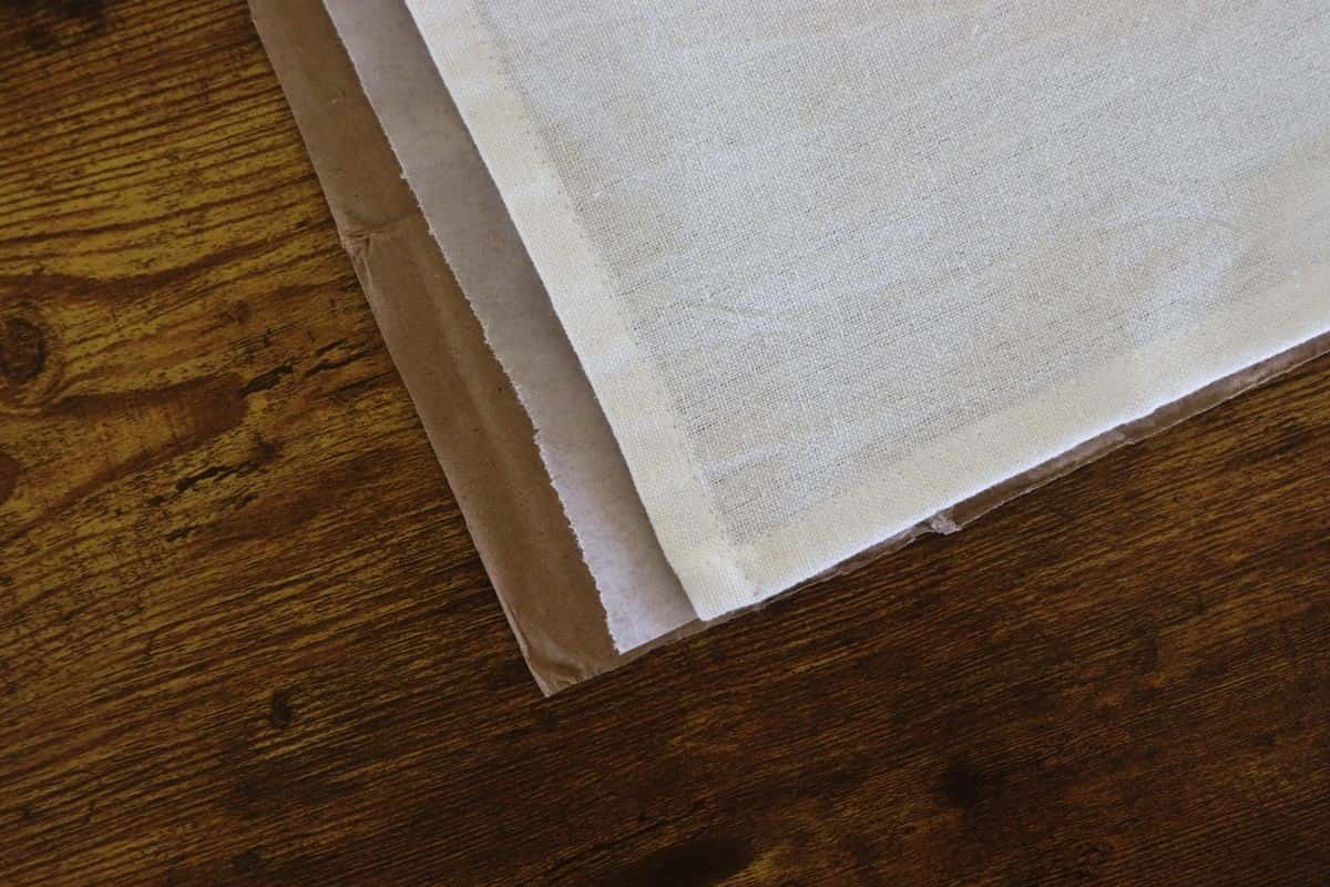 SA piece of cardboard sits on a wooden surface with a pieces of wax paper on top of it and a tea towel on top of that.