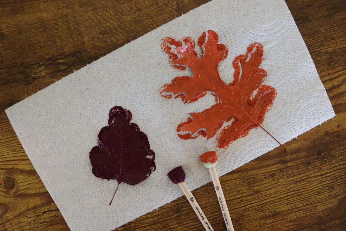A paper towel with a leaf covered in purple paint on it and another leaf covered in orange paint on it. There is a sponge brush next to each leaf with paint on them.