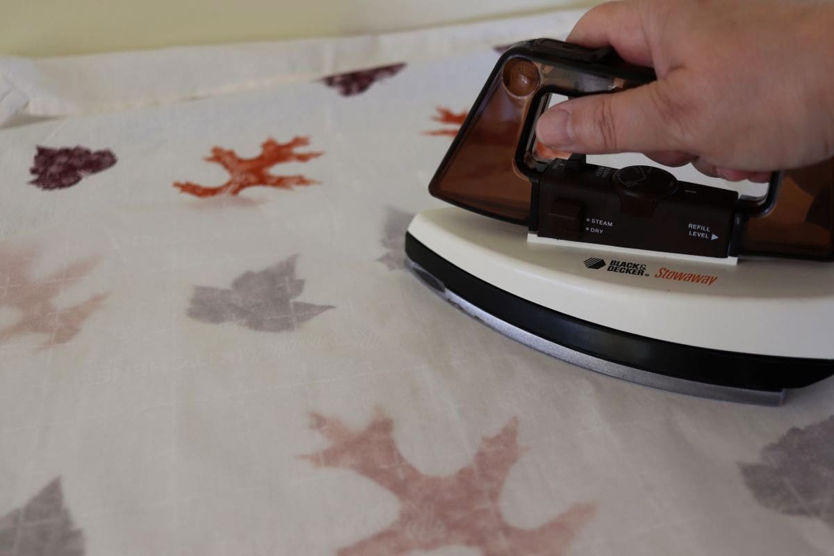 A tea towel with orange and purple leaves on it covered by a piece of wax paper being pressed with an iron.