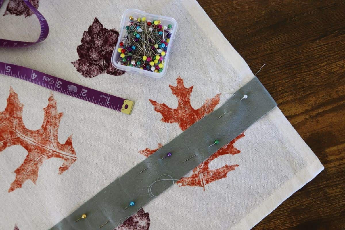 Tea towel with orange and purple leaves on it and a green strip of fabric pinned across the bottom of it. A dish of sewing pins and a measuring tape are sitting on the towel.