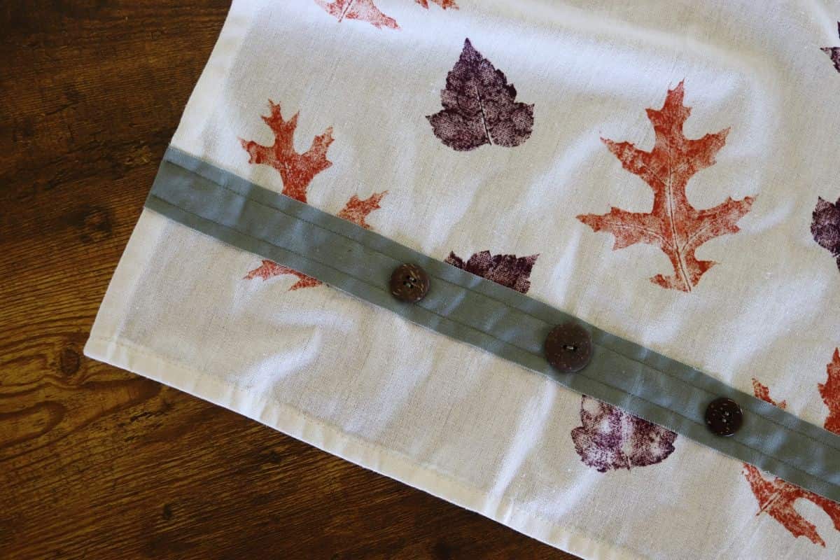Tea towel with orange and purple leaves on it and a green strip of fabric with buttons on it sewn across the bottom.