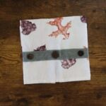 Tea towel with orange and purple leaves on it and a green strip of fabric with buttons across the bottom folded into a square.