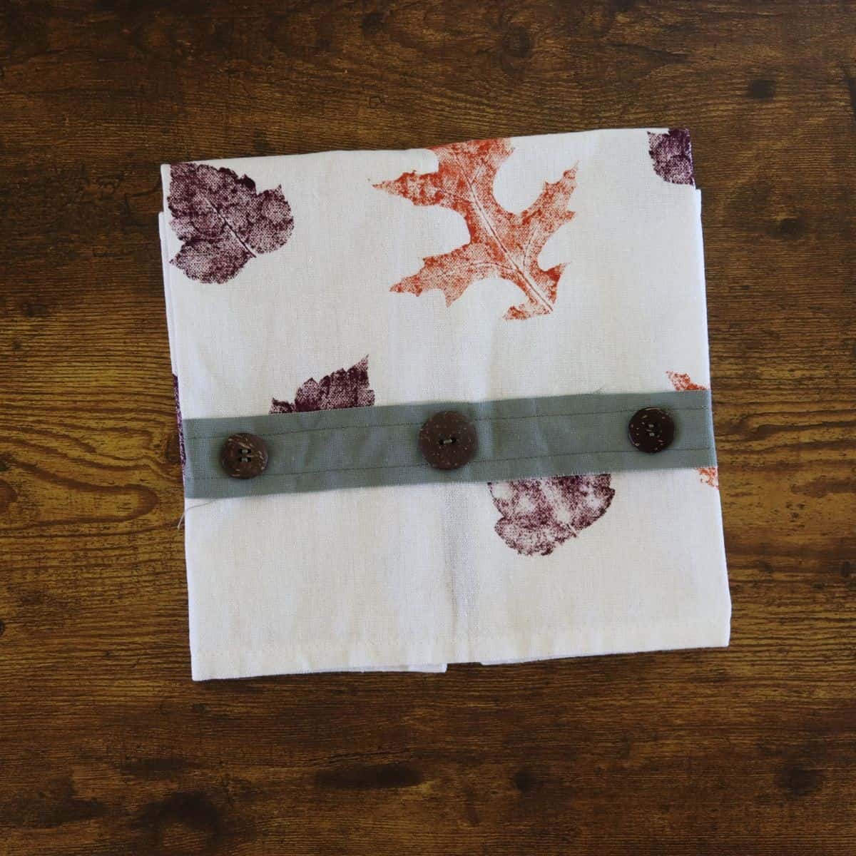 Tea towel with orange and purple leaves on it and a green strip of fabric with buttons across the bottom folded into a square.