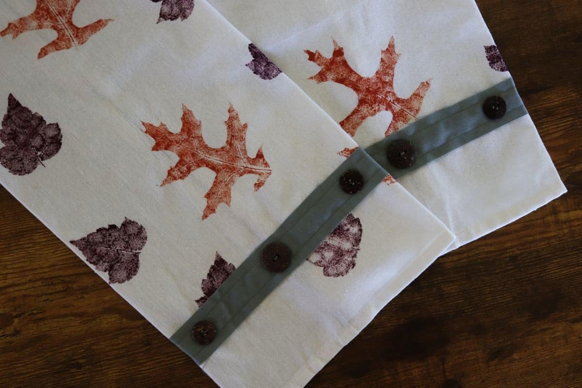 Two white tea towels with stamped leaf patterns in orange and purple tones, each featuring a green strip with brown buttons sewn along one edge, displayed on a wooden surface.