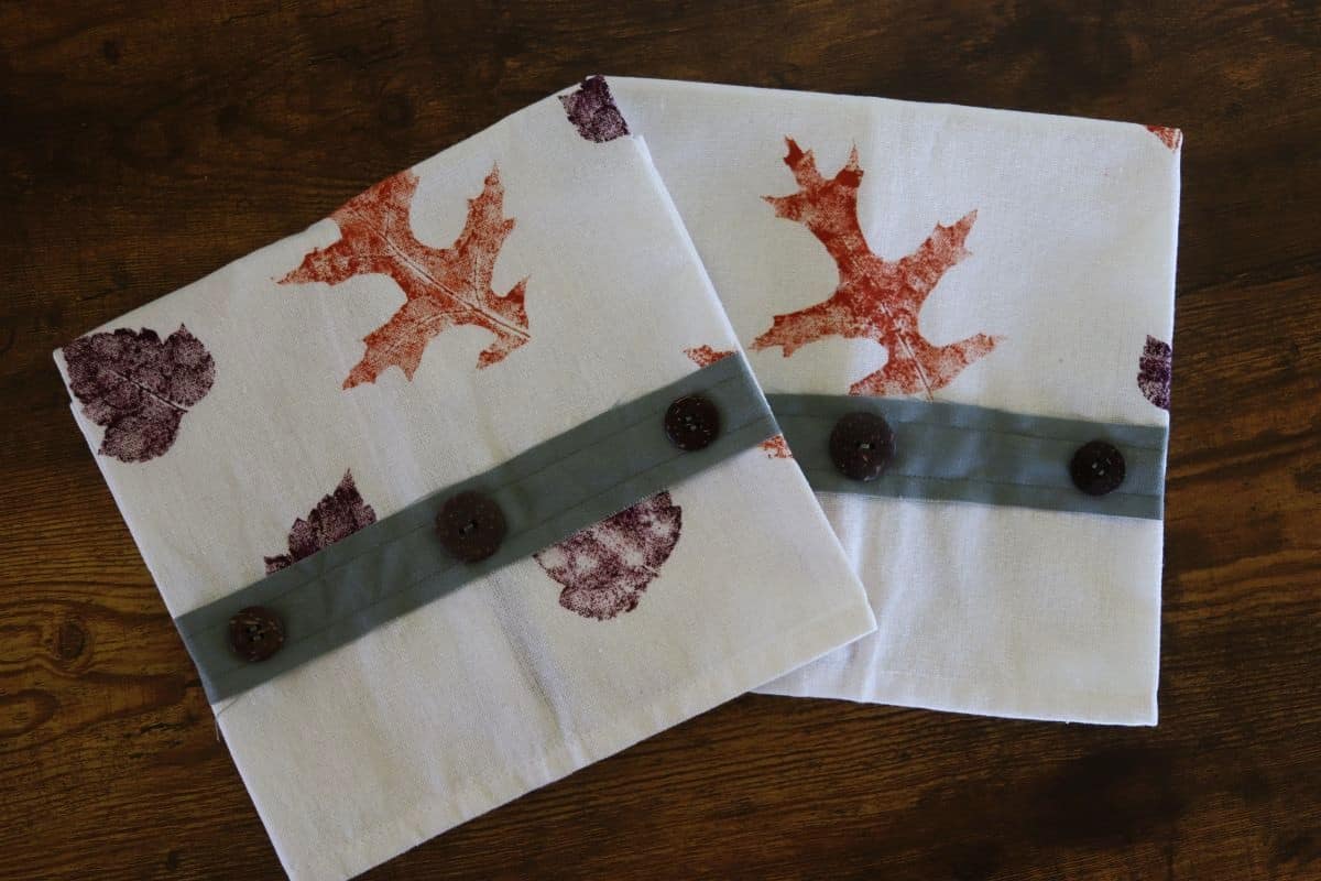 Two white tea towels with stamped leaf patterns in orange and purple. Each towel has a horizontal green strip near one edge, decorated with three dark buttons. They are placed on a wooden surface.