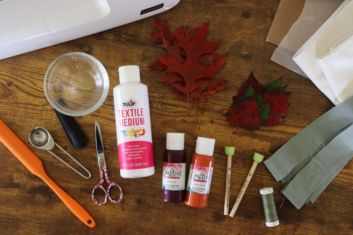 Craft supplies on a wooden table, including fabric paint, textile medium, paintbrushes, scissors, fabric pieces, fresh leaves, a glass bowl, and part of a sewing machine.