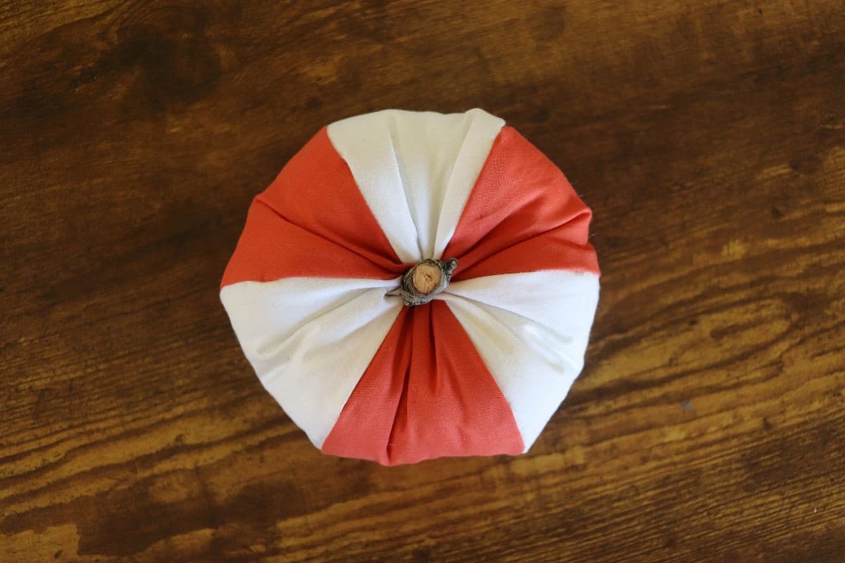 Orange and white fabric shaped pumpkin with a stick stuck in the top opening for a stem.