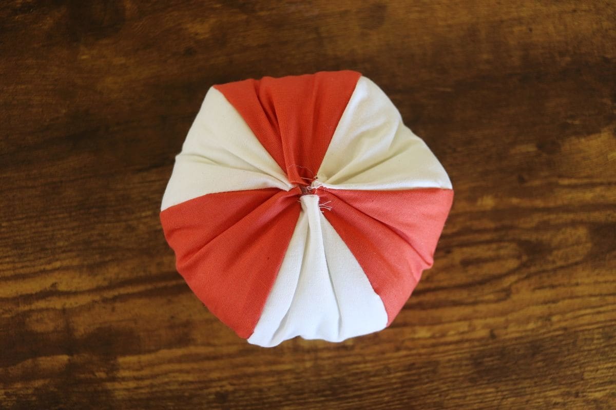 Stuffed ball shape made with orange and white fabric panels sitting on a wooden surface showing the gathering of the opening on the top.