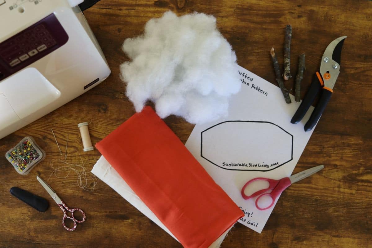 A sewing project setup on a wooden table with orange and cream fabric, sewing supplies, printed pattern, stuffing, twigs, pruners, and a sewing machine. Scissors, pins, and thread are also visible.