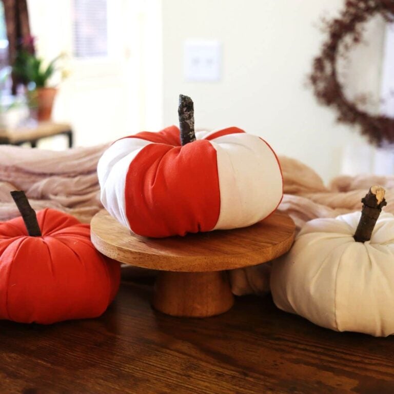 Three stuffed pumpkins, one orange, one white, and one orange and white striped. The striped one sits on a wooden pedestal and the others sit on either side of it.