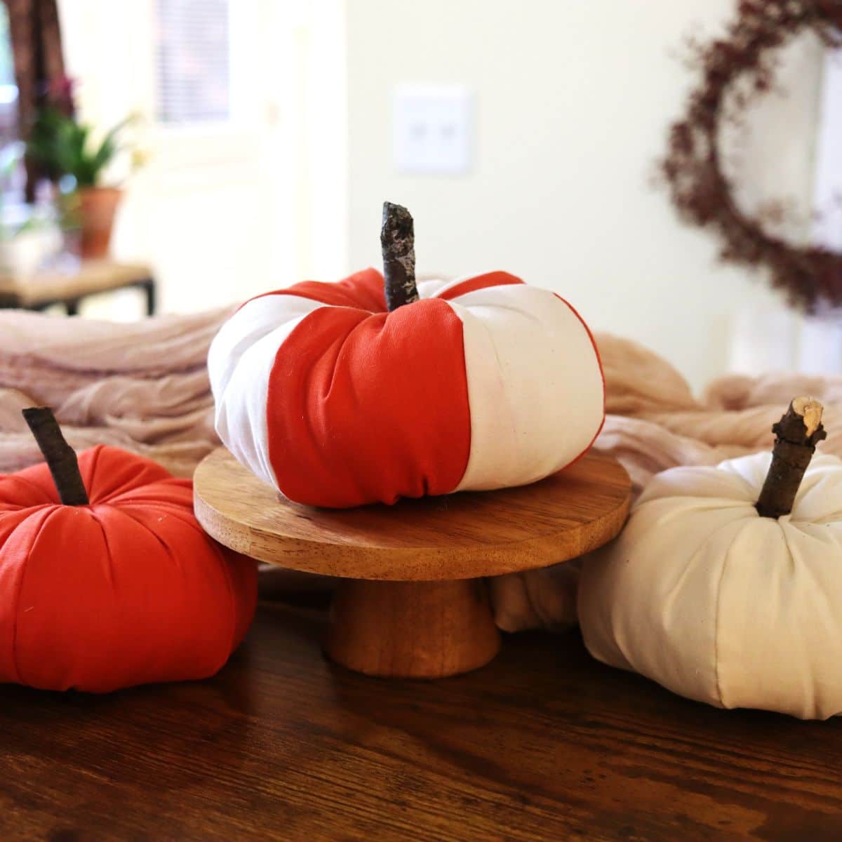 Three stuffed pumpkins, one orange, one white, and one orange and white striped. The striped one sits on a wooden pedestal and the others sit on either side of it.