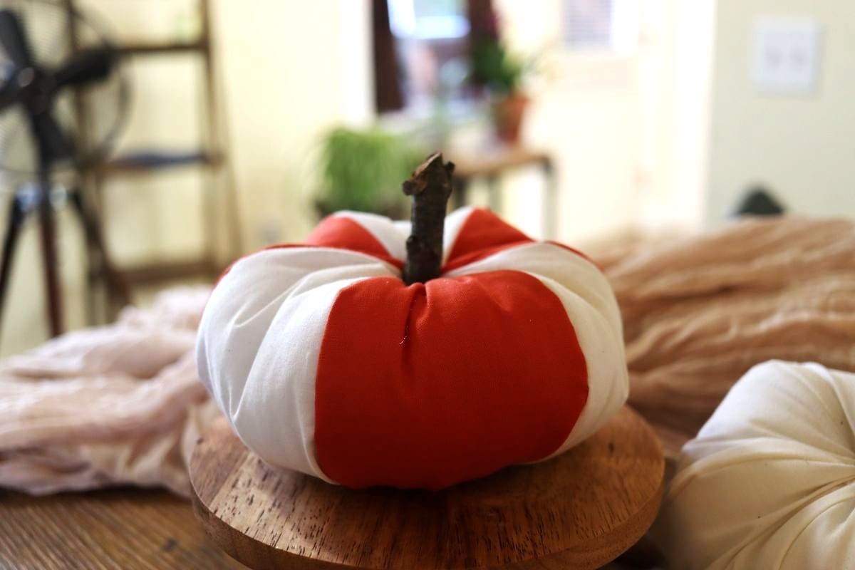 A white and orange fabric pumpkin with a stick stem sits on a round wooden tray, with soft neutral fabric and plants in the blurred background.