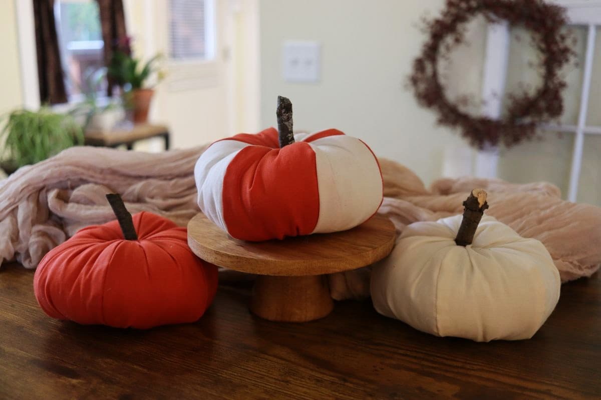 Three fabric pumpkins, one orange, one white, and one with orange and white stripes, sit on a wooden table. One pumpkin is on a wooden stand, all with stick stems. A wreath and plants are visible in the background.