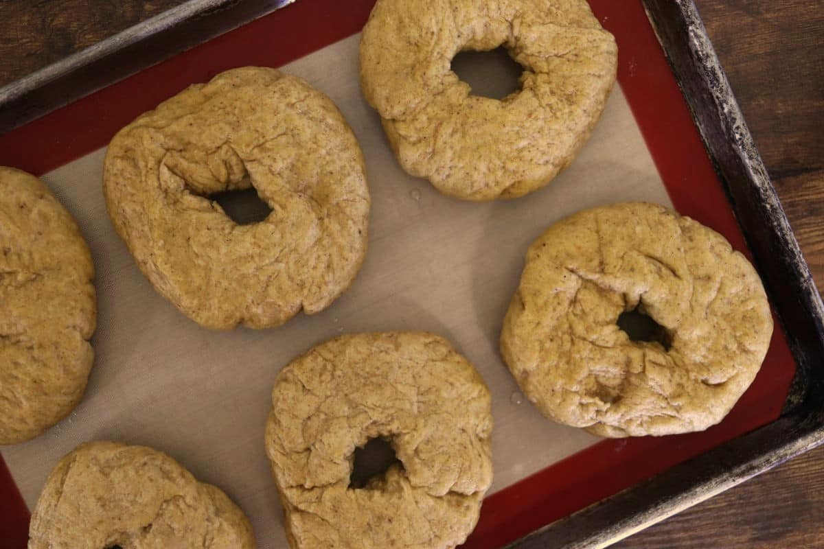 Raw, orange hued bagels on a silicone lined baking sheet.