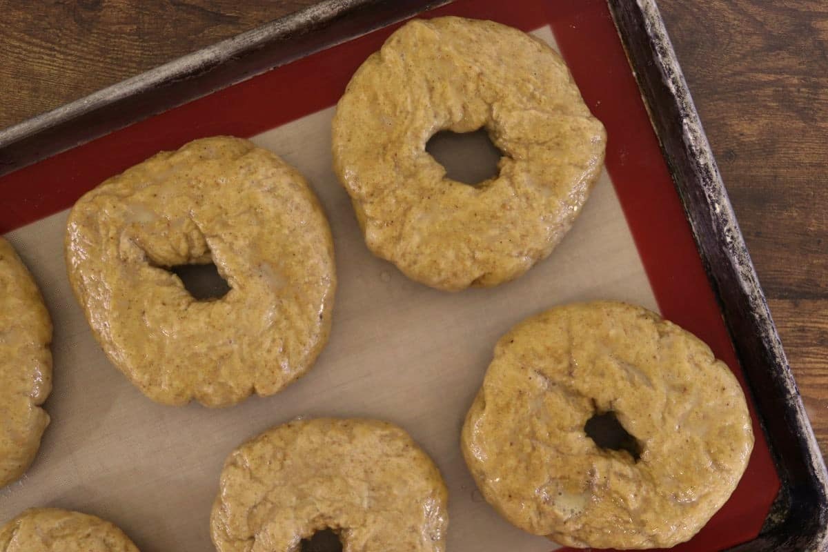 Raw, orange hued bagels, with a wet look on a silicone lined baking sheet.