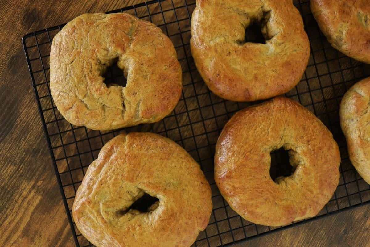 Handmade pumpkin bagels sitting on a black cooling rack on a wooden surface.