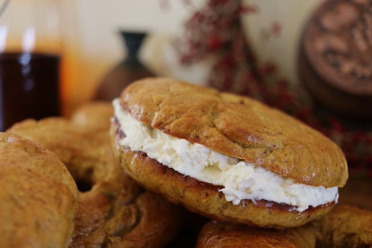 A close-up of a pumpkin bagel with a thick layer of cream cheese filling, surrounded by other bagels. Decorative objects and a blurred background are visible behind them.