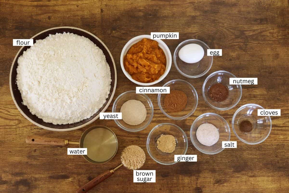 Various labeled baking ingredients are arranged on a wooden surface, including flour, pumpkin puree, an egg, cinnamon, nutmeg, cloves, yeast, ginger, salt, water, and brown sugar in bowls and measuring cups.